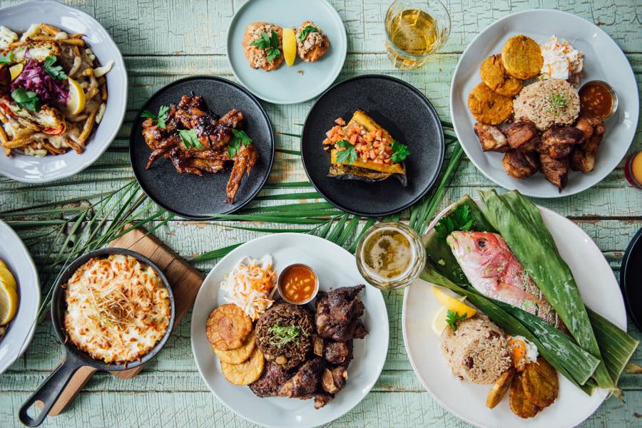 Plates and skillets of Caribbean food on a green wooden table with palm fronds.