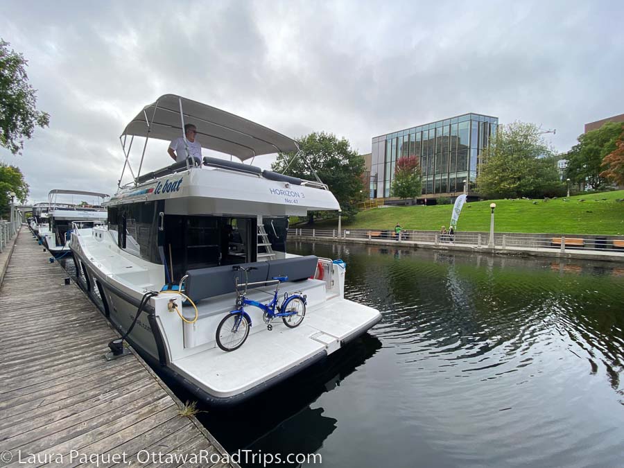 two level white houseboat with a folding bike on the back deck, opposite a large glass building.