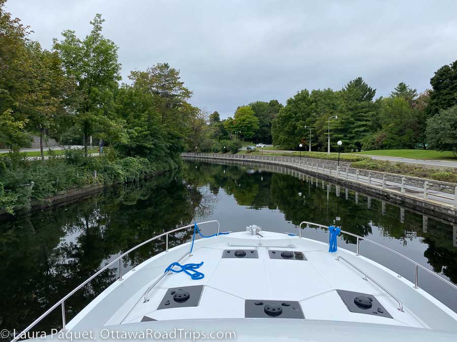 view of the rideau canal over the white bow of a boat, with railings and bike paths on the canal's edges.