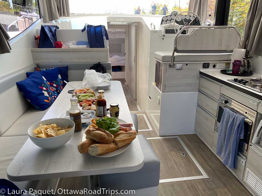 buns, cold cuts and chips on a long table in a galley kitchen, with stove and steering wheel to the right.
