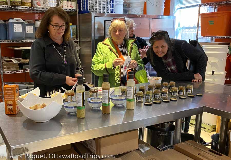 Three women looking at jars of mustard and bottles of dressing on a stainless steel table.