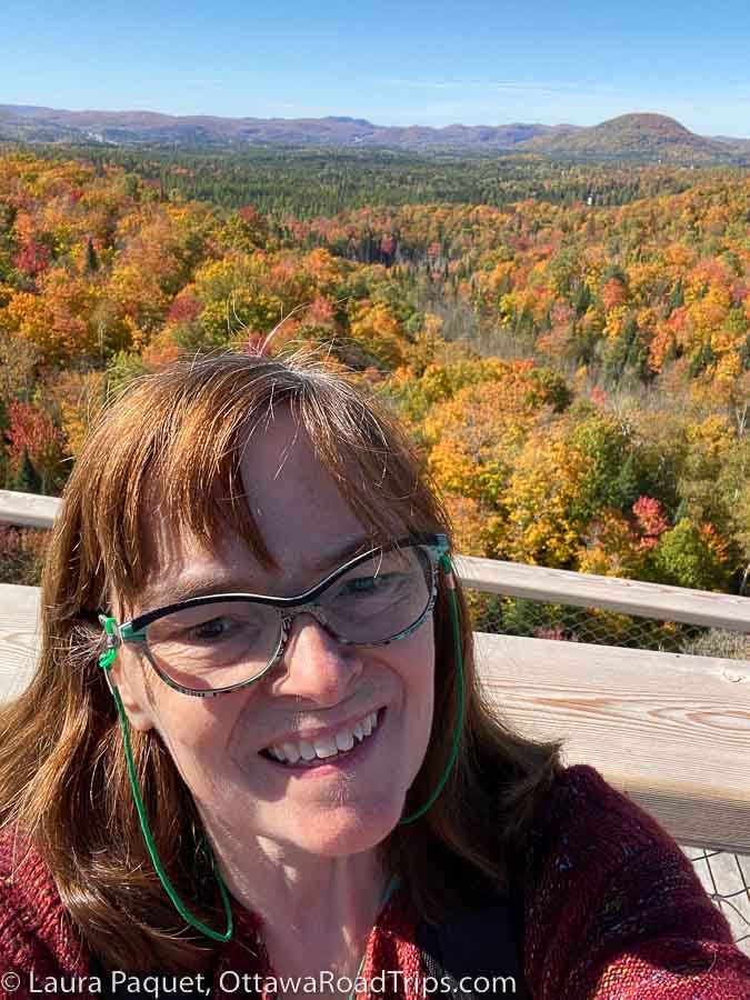 woman with glasses standing on wooden deck, with fall foliage in background.