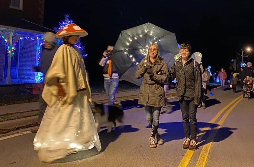 Woman in illuminated Victorian costume, woman with illuminated umbrella and woman wearing illuminated wings, on a paved road.