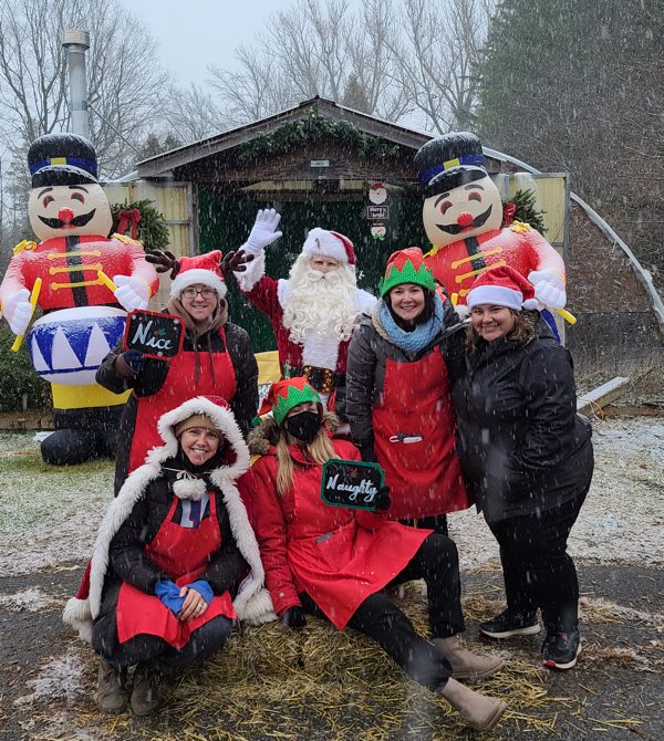 vendors wearing red aprons and elf hats, with santa and two inflated toy soldiers, outside a shed in the snow at kemptville outdoor holiday market