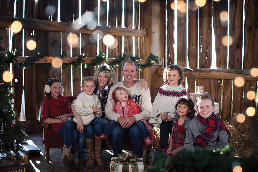 Family with parents and six children in Christmas sweaters and scarves, inside a barn decorated for Christmas.