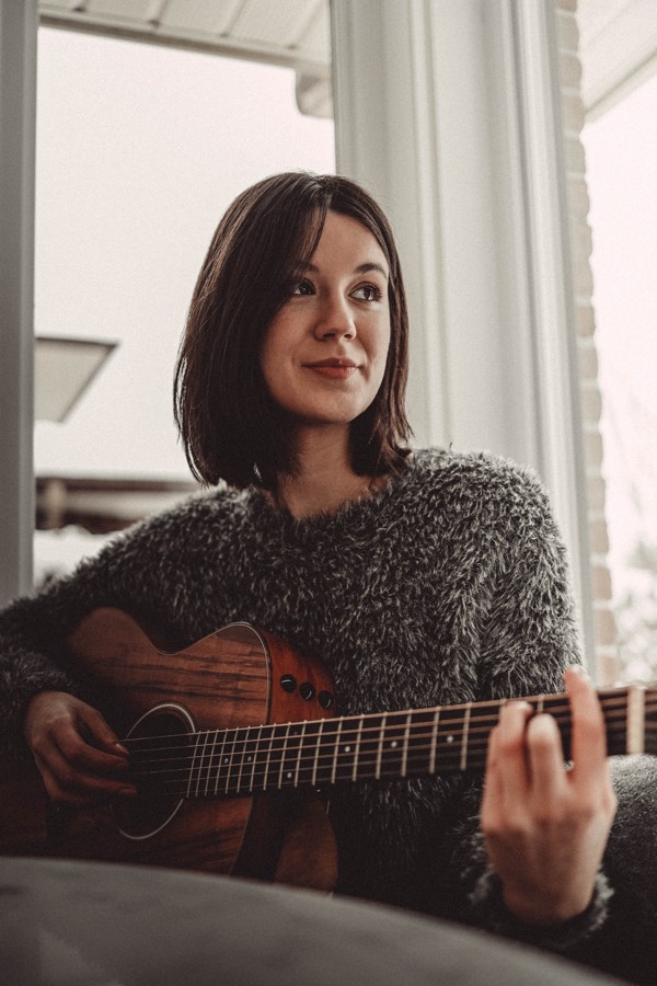 Dark-haired young woman in grey sweater playing an acoustic guitar.