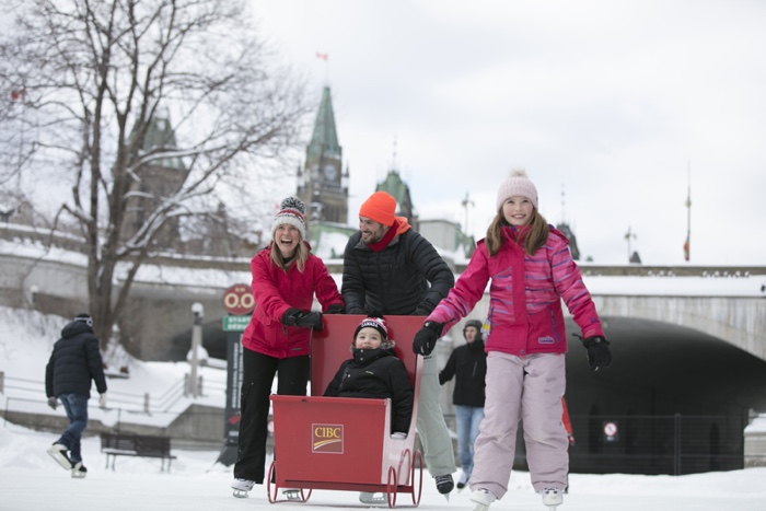 mom and dad with child in red sled and older girl on skates on rideau canal with parliament hill in background.