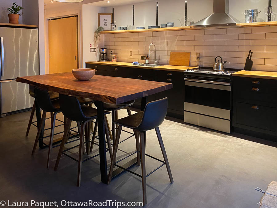 kitchen with black cabinets, stainless-steel oven and fridge, wooden table with six bar stools, and concrete floor.