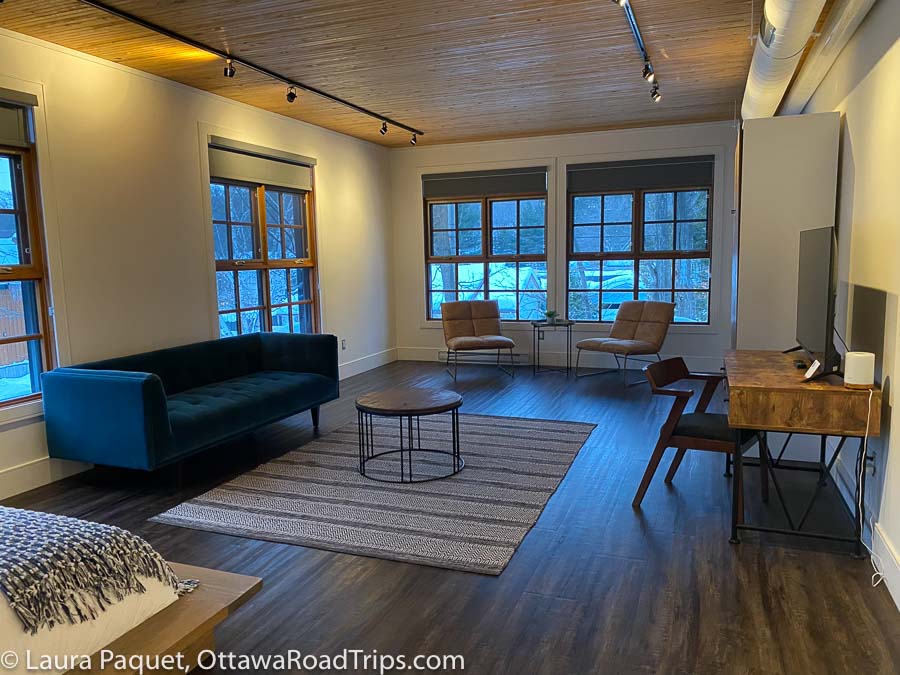 room with large windows, blue modern couch, two leather chairs, murphy bed cabinet, desk with tv, wood floors