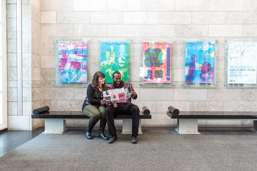 couple sitting on a bench reading a paper guide with canadian centre for architecture posters on wall behind