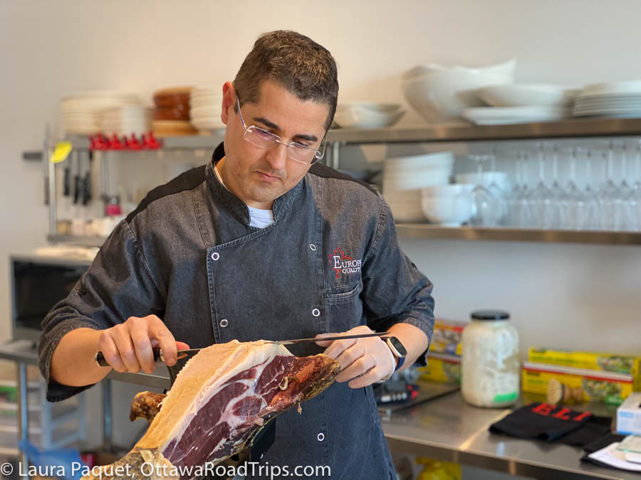 man in denim chef's coat carving large piece of Joselito ham at El Rebost de les Mil Iles in Prescott, Ontario.