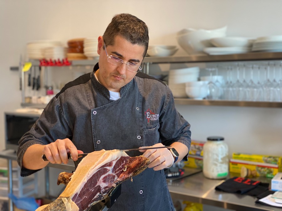 man in denim chef's coat carving thin slices from a large cured ham