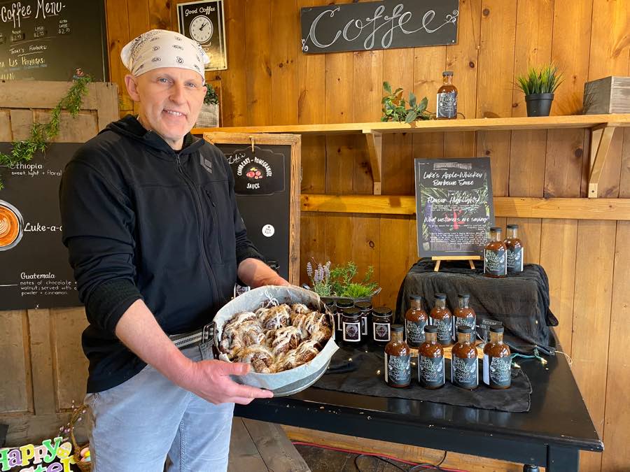 man in black sweatshirt holding a pan of cinnamon buns in front a display of barbecue sauce