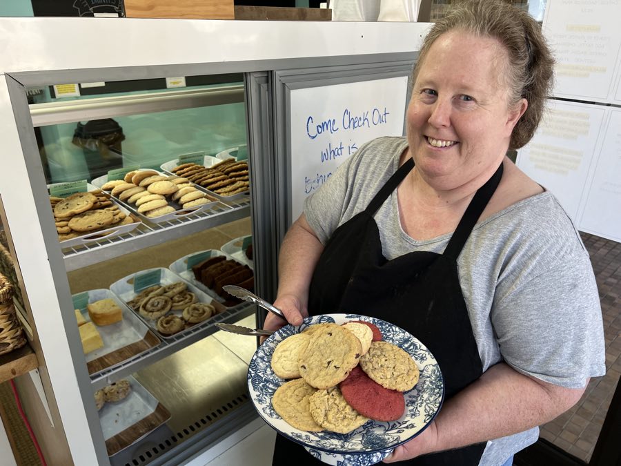 woman in black apron in front of a bakery display case, holding a plate of cookies