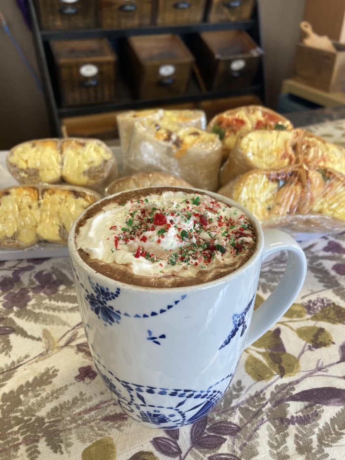 a mug of hot chocolate with red and green sprinkles and whipped cream, with wrapped sandwiches in background