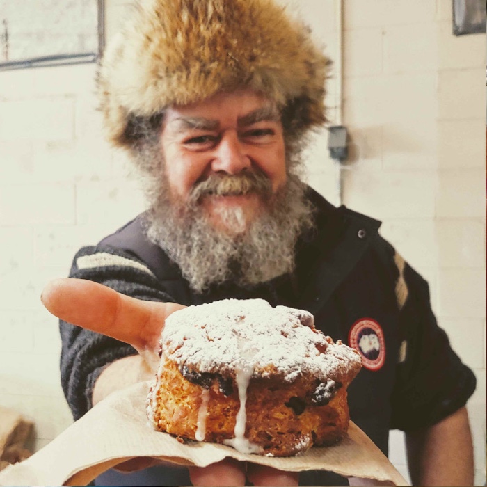 bearded man with fuzzy hat holding a scone toward the camera