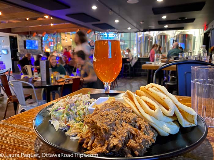 round black plate with pieces of naan, a mound of pulled pork and a scoop of coleslaw, with a glass of beer in background, at the big slide brewery and public house in lake placid