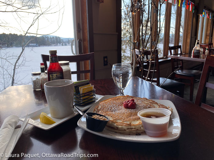 square white plate of pancakes with raspberries, maple syrup and cinnamon butter, with snow-covered lake in background