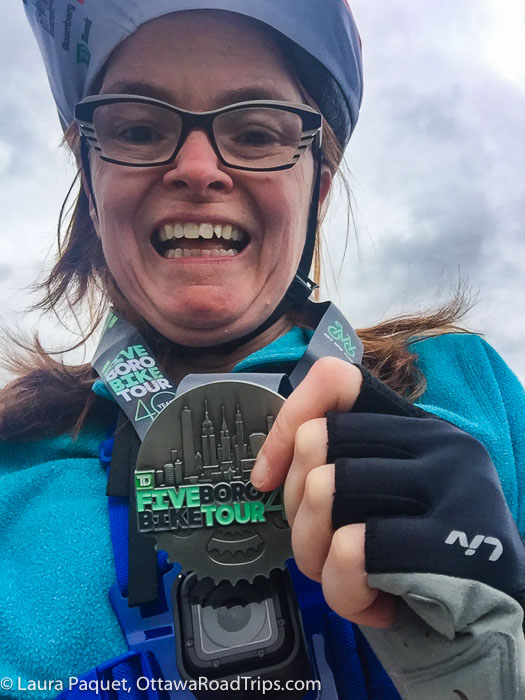 smiling woman holding a pewter five boro bike tour medal on a ribbon around her neck