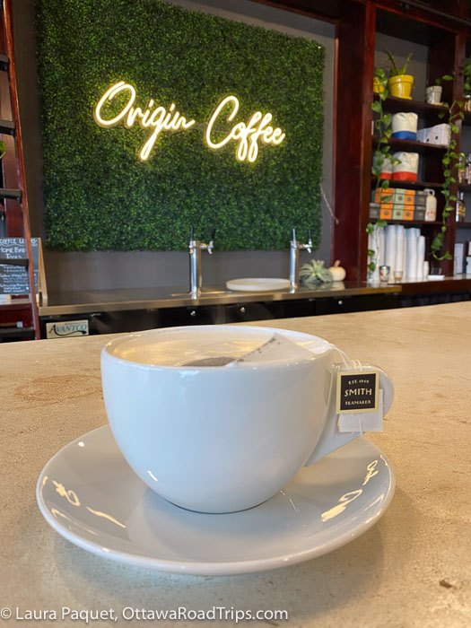 closeup of a large white teacup filled with foamy tea, with shelves of coffee and tea in background, at origin coffee co. in lake placid