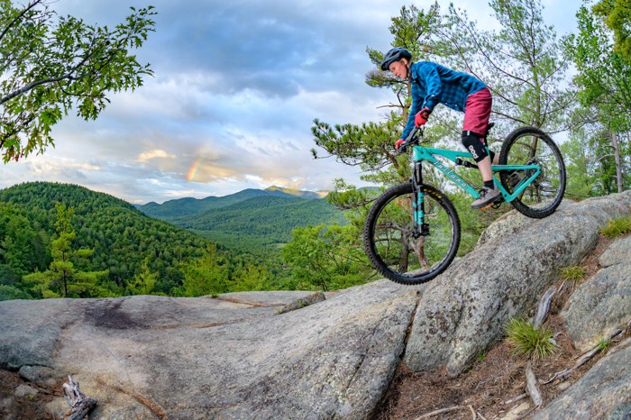 person on a blue mountain bike pedalling over rocks with green mountains in background