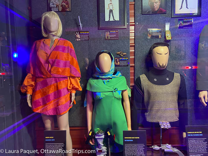 mannequins in a glass display case wearing an orange-and-pink dress, a green dress, and a klingon costume
