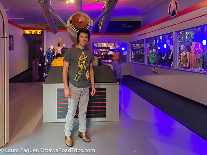 man in t-shirt and jeans in front of a large model of the uss enterprise and display cases of star trek memorabilia