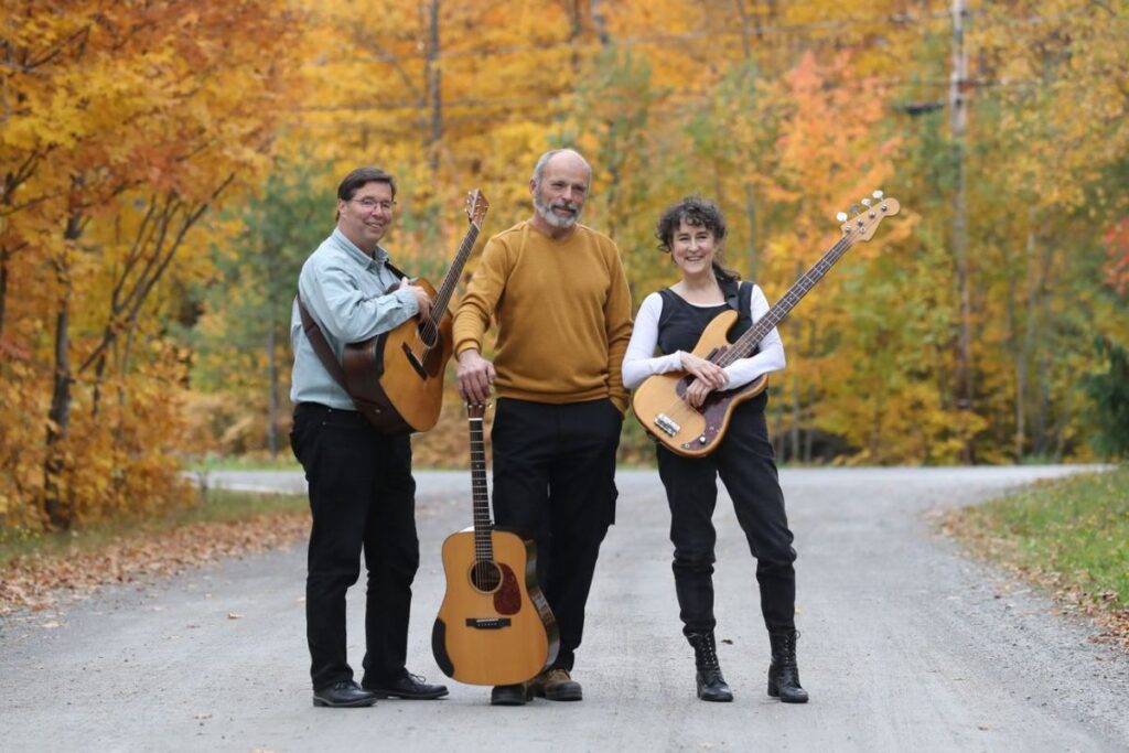 two men with acoustic guitars and a woman with an electric car standing on a gravel country road in fall