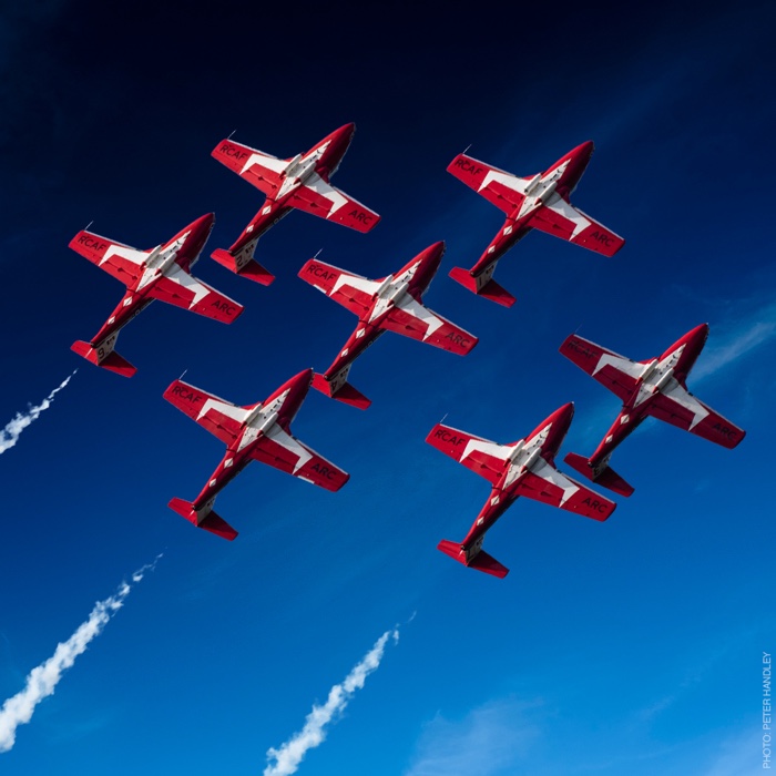 seven red-and-white snowbirds planes flying in formation against a blue sky.