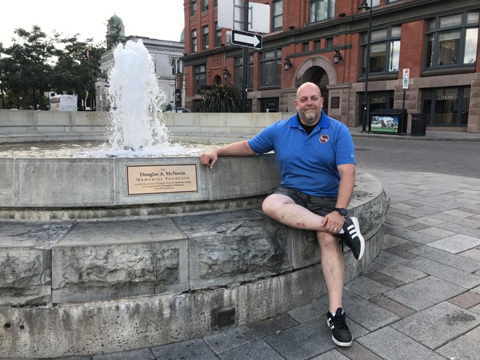 bearded man in blue polo shirt and shorts sitting next to a fountain.