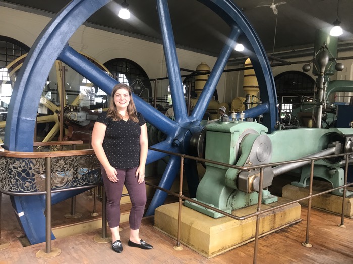 woman standing in front of a large blue mechanical wheel at the pumphouse museum in kingston.