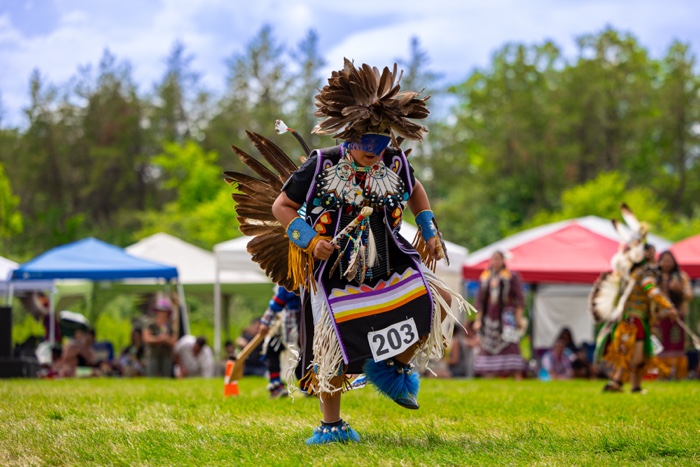 man in pow wow regalia dancing on grass with awnings and other dancers in background