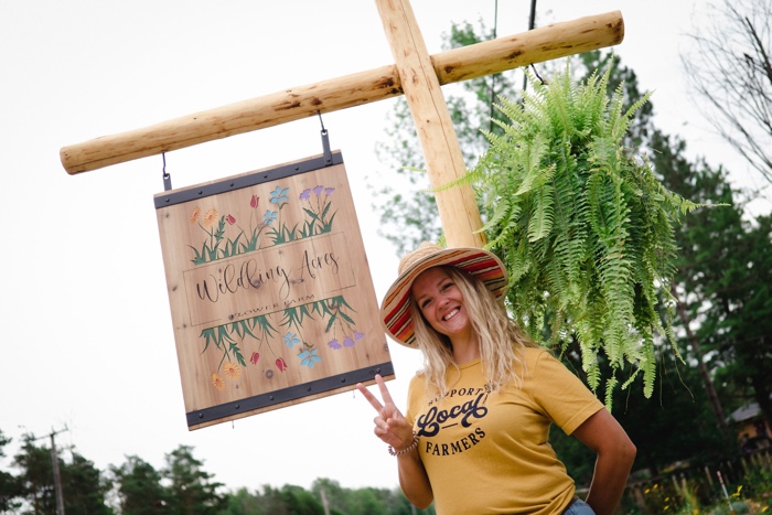 woman smiling and making a peace sign under a wooden sign reading wilding acres.