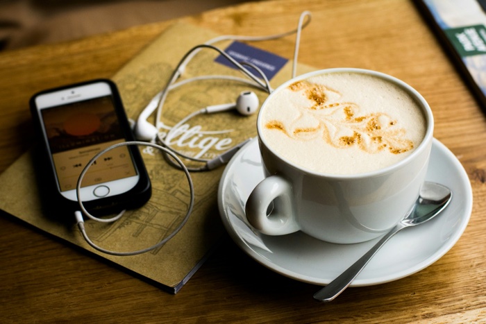 cellphone and earphones next to a white cup of café au lait on a wooden table