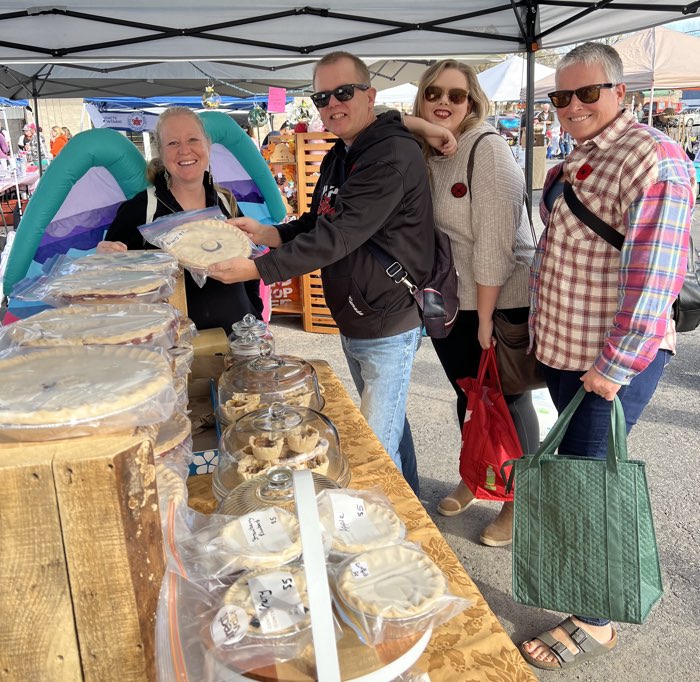 four people under a white awning at a bakery stand at a farmers' market, with one person holding a pie