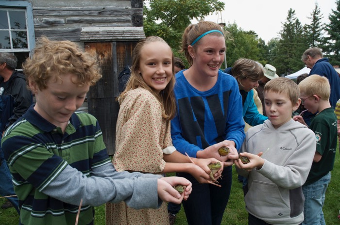 two boys and two girls holding frogs in front of a wooden building at the glengarry pioneer museum in dunvegan. photo courtesy of the museum.