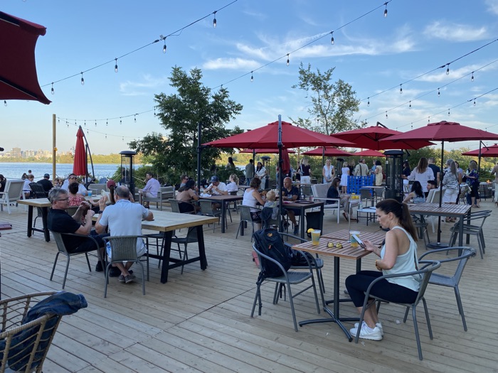 people sitting on metal chairs by wooden tables on deck overlooking the ottawa river at tavern on the island