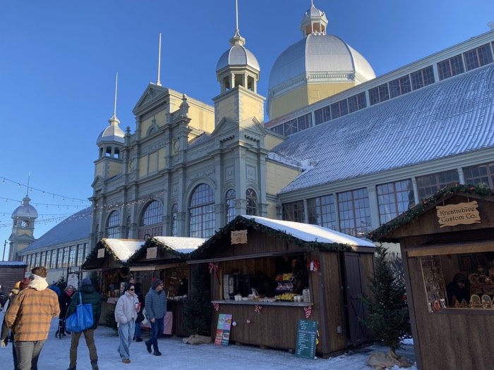 people in winter coats walking by wooden huts selling food and christmas crafts, with domed aberdeen pavilion in background