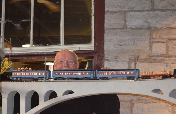 grey-haired man smiling as he stands behind a model train on a bridge