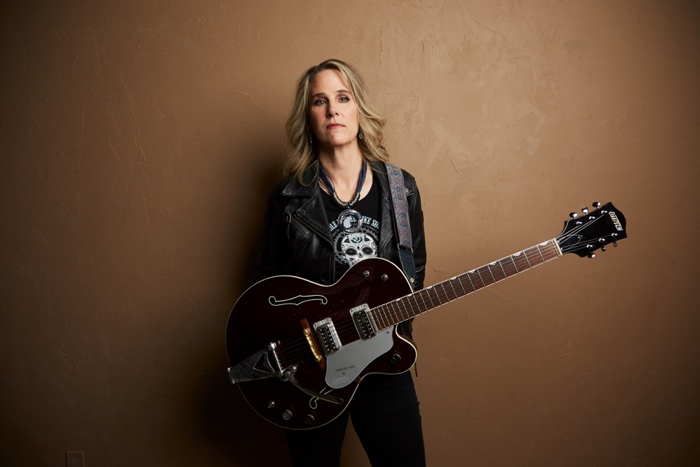 blonde woman in leather jacket with guitar hanging from a strap around her neck