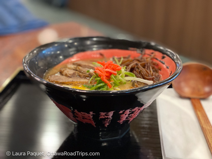 closeup of a black pottery bowl of ramen on a black lacquered tray, with a wooden ladle-type spoon