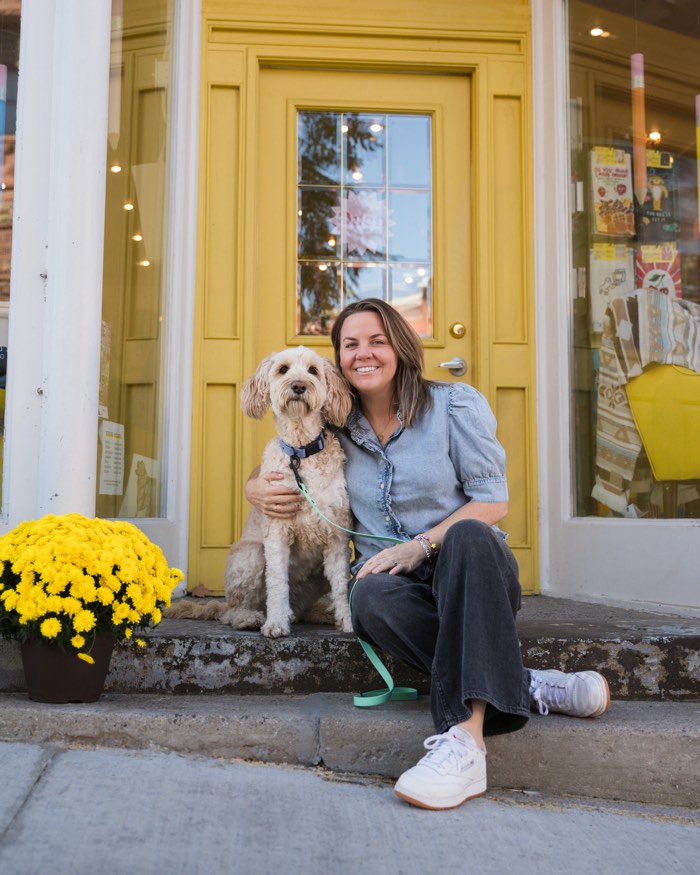 smiling woman sitting with a dog on steps of a shop with a yellow door