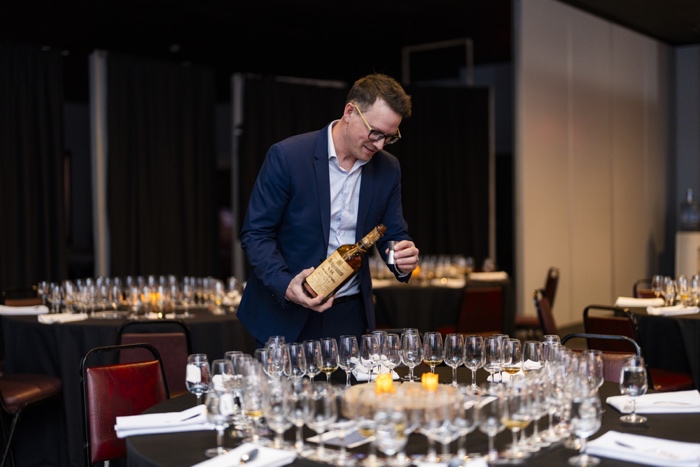 man pouring whiskey into a silver jigger, in front of a large table of glasses