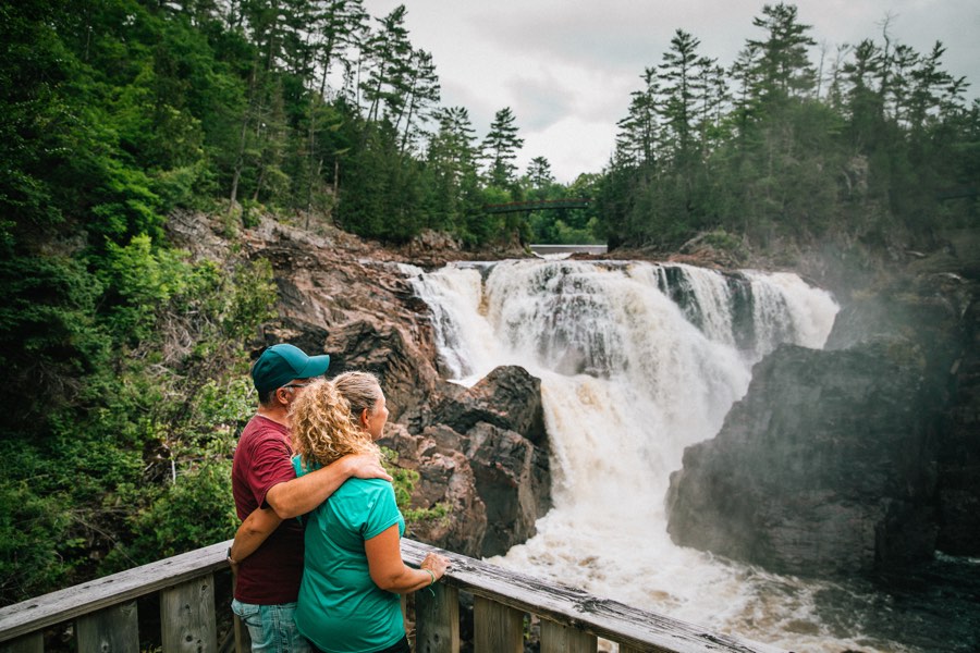 man and woman standing on a wooden platform overlooking a rushing waterfall.