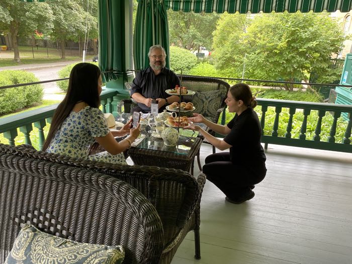 two people sitting on wicker love seats admiring and photographing a tea tray while a server explains what's on it