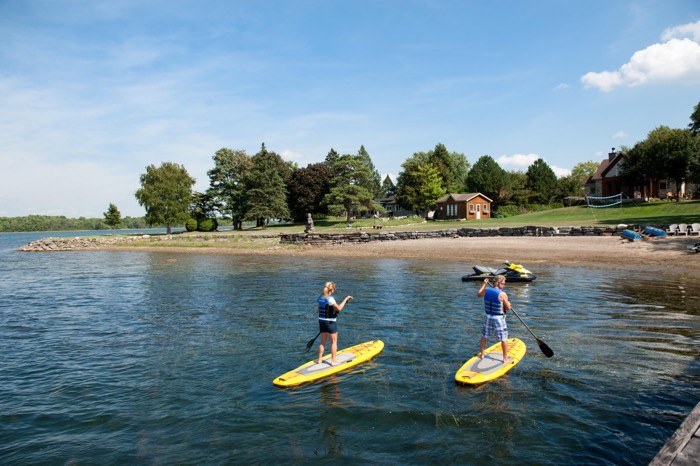 two people on yellow standup paddleboards in the water near a small beach and cottages.