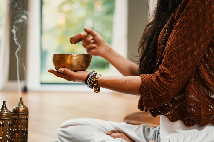 closeup of a woman's hands holding a singing bowl and a stick, with incense burning nearby