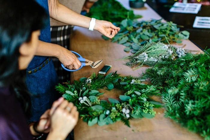 closeup of people's hands as they make green wreaths at a long table