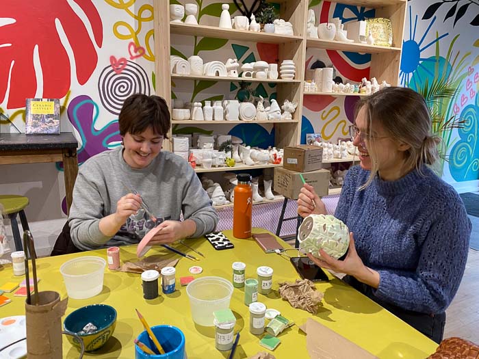 two young women sitting at a yellow table painting pottery