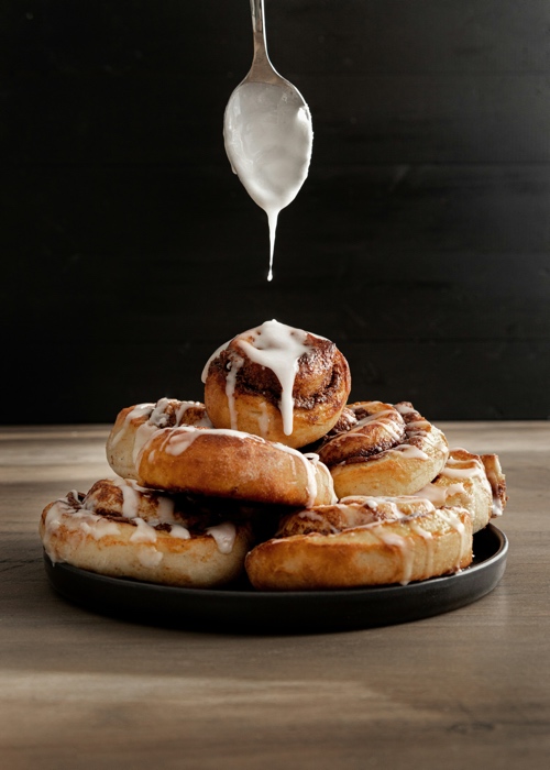 eve-maier-cinnamon rolls-unsplash - Ottawa Road Trips a spoon dripping white icing above a round plate of partly iced cinnamon rolls